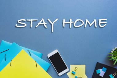 Kırtasiye ve akıllı telefonun üst görünümü ve mavi yüzey üzerinde boş ekran ile evde kalma harfleri