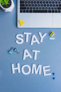 Top view of stay at home lettering near laptop, plant and stationery on blue background clipart