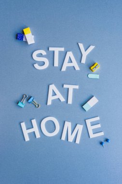 Top view of stay at home lettering near erasers, binder clips and pencil sharpener on blue surface clipart