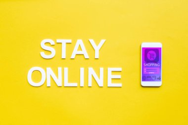 Top view of stay online lettering and smartphone with online shopping app on yellow surface clipart
