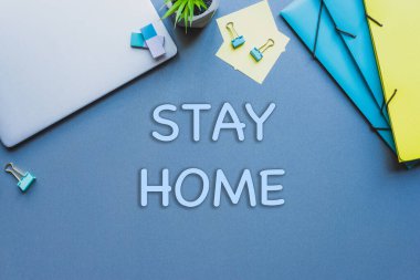 Top view of stay home lettering near laptop, plant and stationery on blue surface clipart