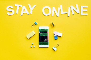 Top view of stay online lettering near smartphone with booking app on yellow surface clipart