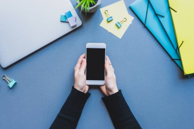 Dizüstü bilgisayarın yanında akıllı telefonu ve mavi yüzeyde kırtasiyeyi tutan kadının en üst görüntüsü