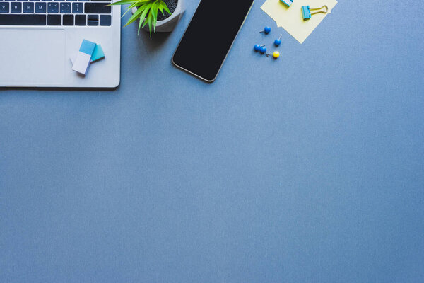 Top view of laptop, smartphone with blank screen and stationery on blue background