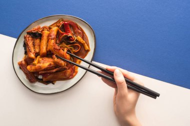cropped view of woman holding chopsticks near korean topokki with sauce on white and blue clipart