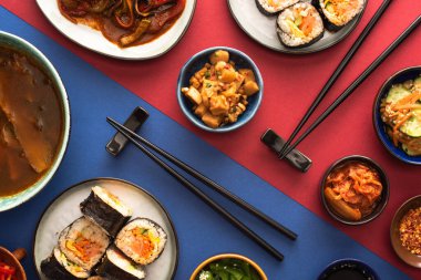 top view of chopsticks near ramen, kimbap and korean side dishes on blue and crimson  clipart