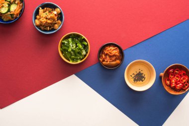 top view of bowls with korean side dishes and oil on blue, crimson and white  clipart