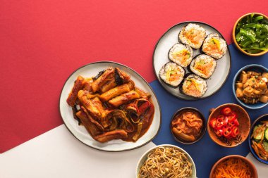 top view of korean topokki near bowls with spicy side dishes on blue, crimson and white  clipart
