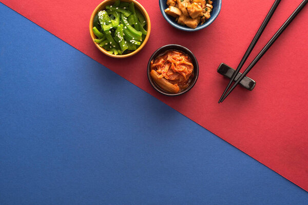 top view of bowls with spicy korean food near chopsticks on blue and crimson  