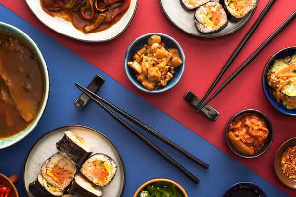 top view of chopsticks near ramen, kimbap and korean side dishes on blue and crimson 