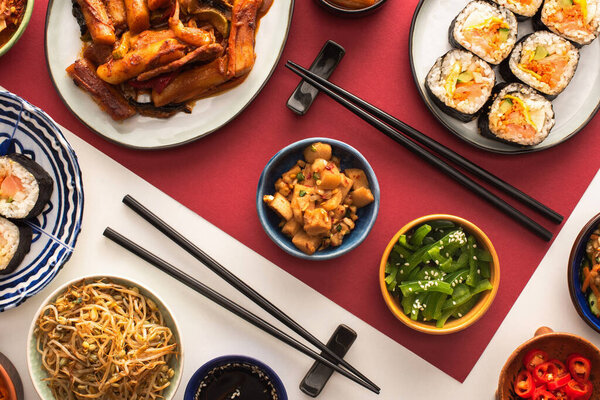 top view of chopsticks near korean topokki and traditional side dishes on white and crimson 