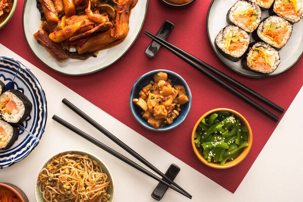 top view of chopsticks near topokki and traditional korean side dishes on white and crimson 