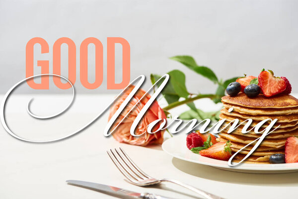 selective focus of delicious pancakes with blueberries and strawberries on plate near rose flower and cutlery on white surface isolated on grey, good morning illustration