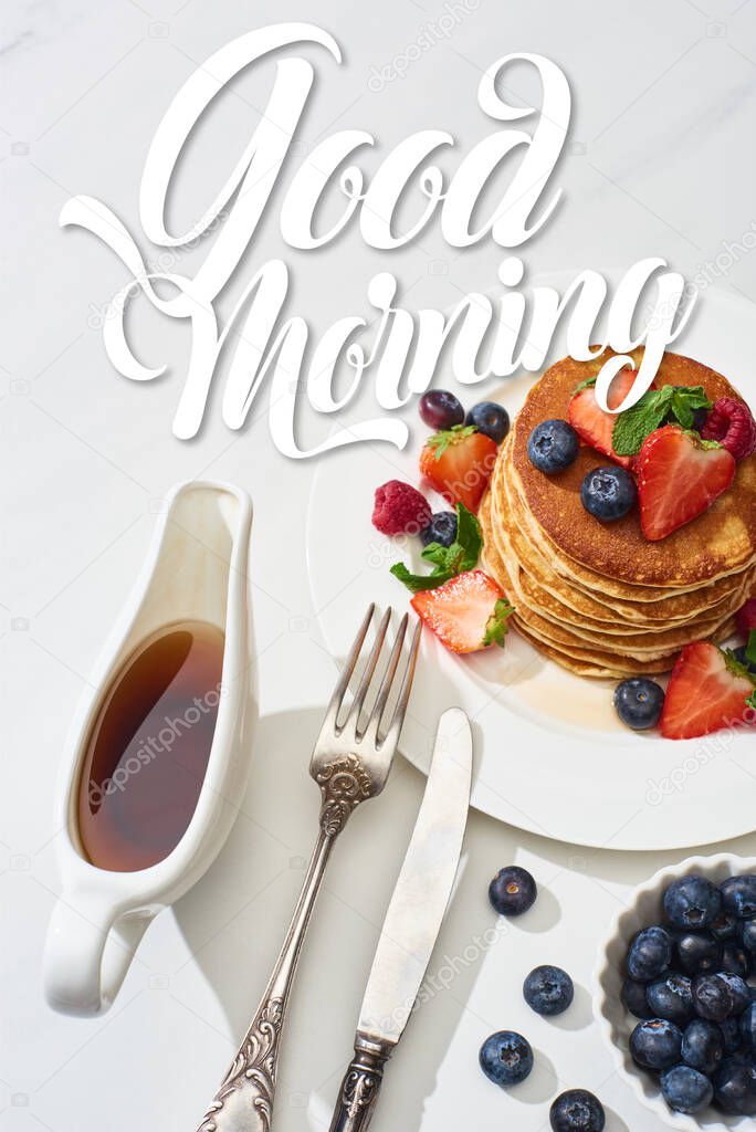 top view of delicious pancakes with maple syrup, blueberries and strawberries on plate near fork and knife on marble white surface, good morning illustration