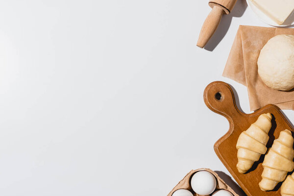 вид на свежие круассаны на деревянной доске возле сырого теста, скалка, масло и яйца на белом фоне
