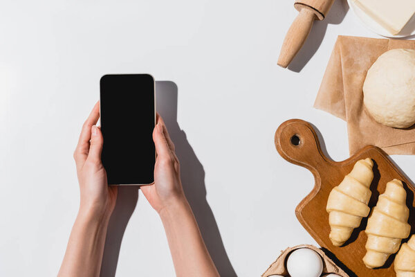 частичный вид женщины, держащей смартфон рядом с сырыми круассанами и ингредиентами на белом фоне
