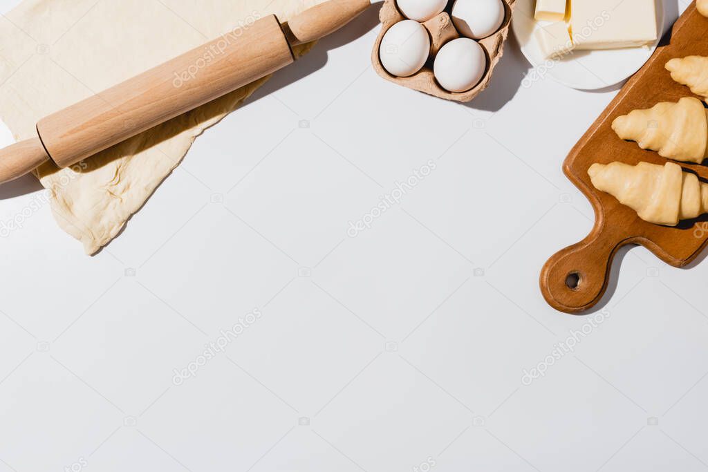 top view of fresh raw croissants on wooden cutting board near rolling pin, butter and eggs on white background