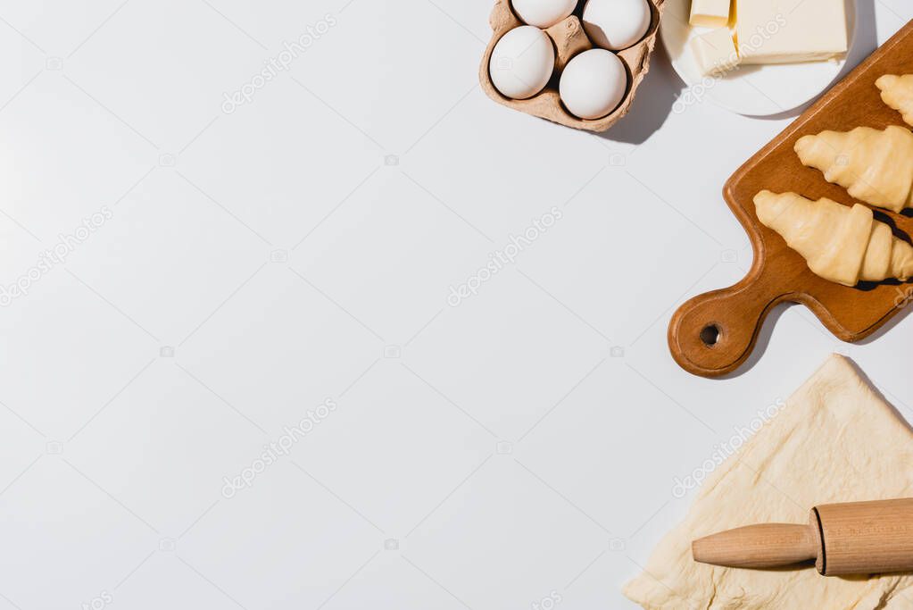 top view of fresh raw croissants on wooden cutting board near rolling pin, butter and eggs on white background