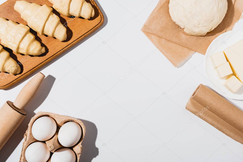 top view of fresh croissants on wooden cutting board near raw dough, rolling pin, butter and eggs on white background