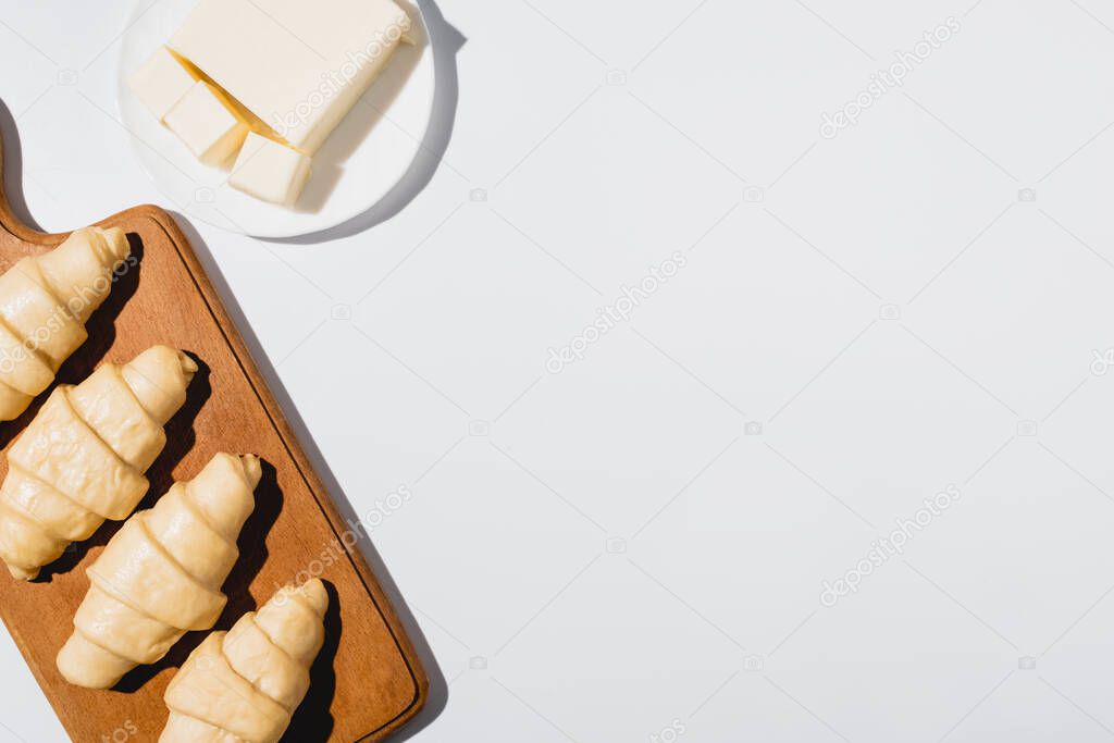 top view of fresh raw croissants on wooden cutting board near butter on plate on white background