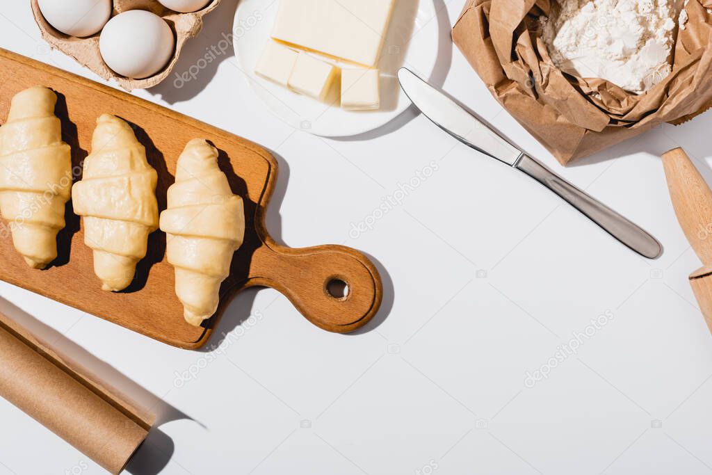 top view of raw croissants on wooden cutting board near ingredients on white background