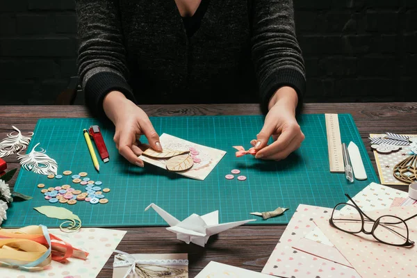 Cropped image of woman adding bow to handmade scrapbooking postcard — Stock Photo