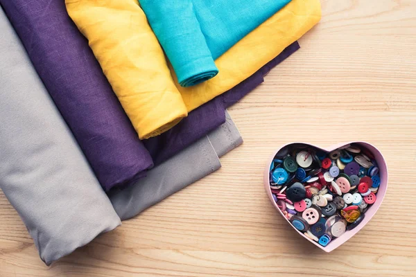 Top view of wooden table with rolls of fabric and heart shaped box with buttons — Stock Photo