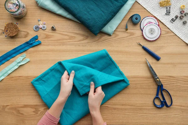 Vista superior do tecido dobrável das mãos femininas cortadas no local de trabalho da costureira com tesouras e botões — Fotografia de Stock