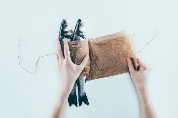 Tiro recortado de manos envolviendo peces de mar en papel y atando con cuerda en gris - foto de stock