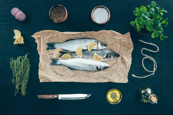 Vue de dessus du poisson gastronomique avec des tranches de citron sur papier cuisson et des épices sur noir — Photo de stock