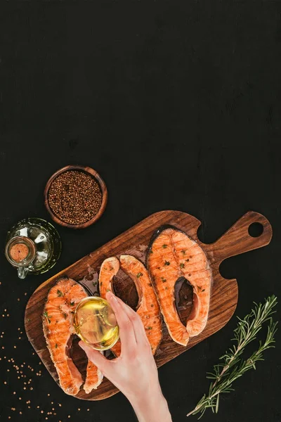 Top view of hand pouring olive oil on delicious salmon steaks with spices on black — Stock Photo