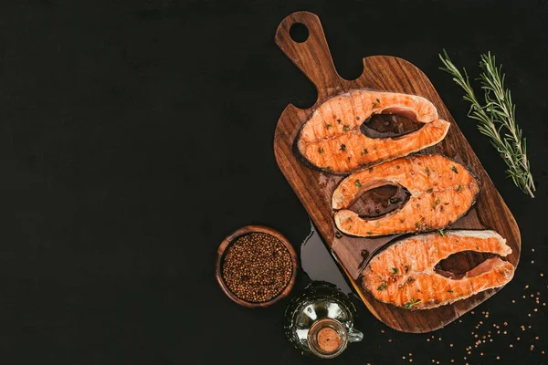 Vista dall'alto di bistecche di salmone gourmet su tagliere di legno con spezie su nero — Foto stock