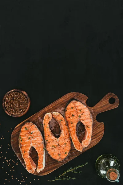 Top view of salmon steaks on wooden cutting board with spices on black — Stock Photo