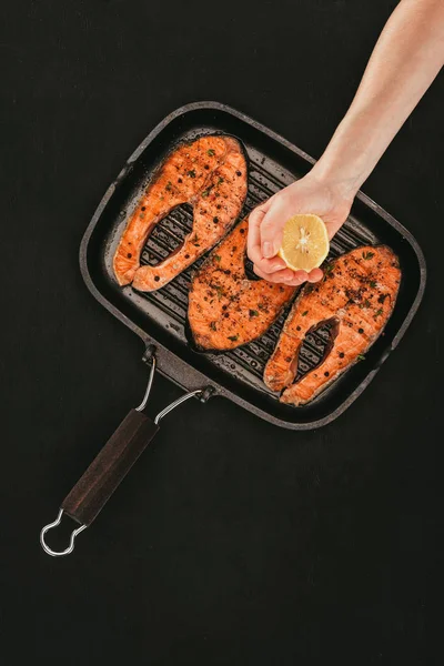 Cropped shot of person squeezing lemon juice above salmon steaks on grill on black — Stock Photo