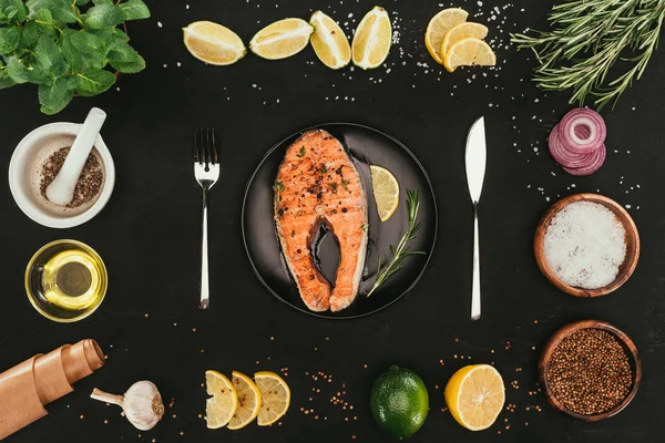 Top view of salmon steak with cutlery and seasonings on black — Stock Photo