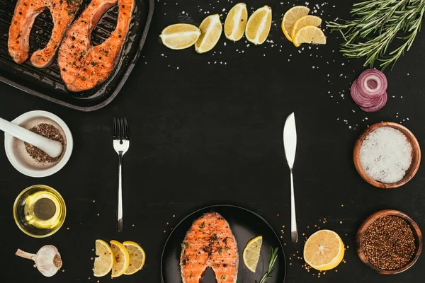 Top view of grilled salmon steaks, cutlery and spices on black — Stock Photo