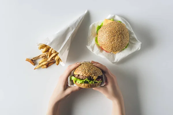 Vista recortada de las manos con hamburguesas y papas fritas en cono de papel, aislado en blanco - foto de stock