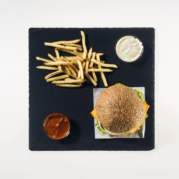 Vista superior de hambúrguer, batatas fritas e molhos em placa de pedra preta, isolado em branco — Fotografia de Stock