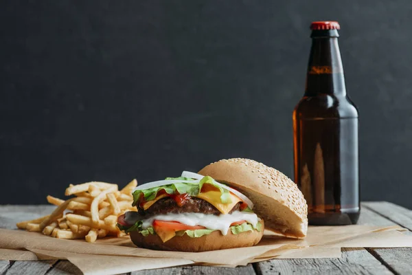 Traditional homemade hamburger, french fries and bottle of beer on baking paper on wooden tabletop — Stock Photo