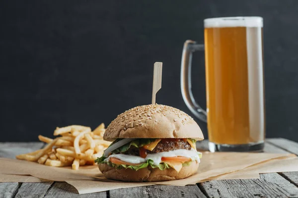 Hamburguesa casera tradicional, papas fritas y un vaso de cerveza sobre papel de hornear sobre mesa de madera - foto de stock