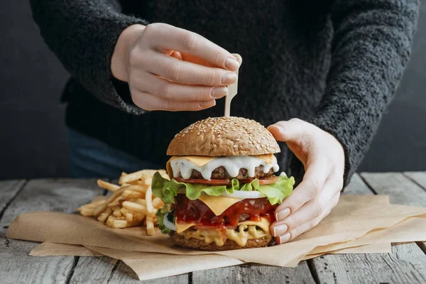 Vista recortada de las manos femeninas con una gran hamburguesa con queso y papas fritas en papel para hornear - foto de stock