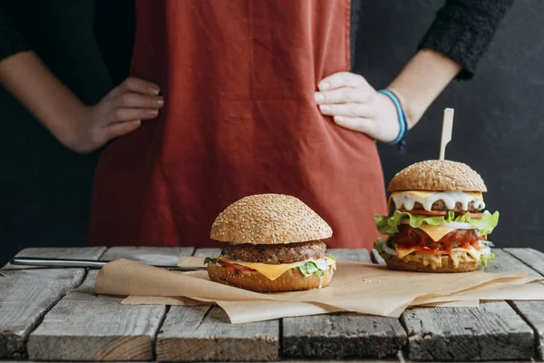 Cropped view of girl in apron standing at wooden table with homemade cheeseburgers on baking paper — Stock Photo