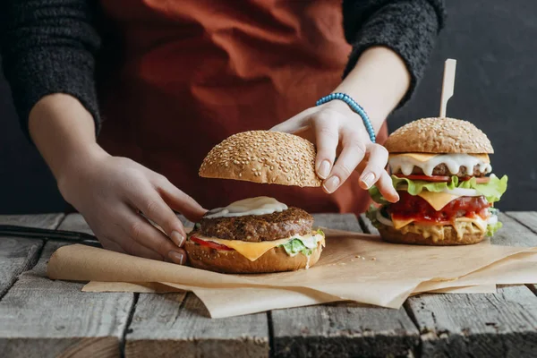 Abgeschnittene Ansicht von Mädchen in Schürze machen hausgemachte Cheeseburger auf Backpapier auf hölzerner Tischplatte — Stockfoto