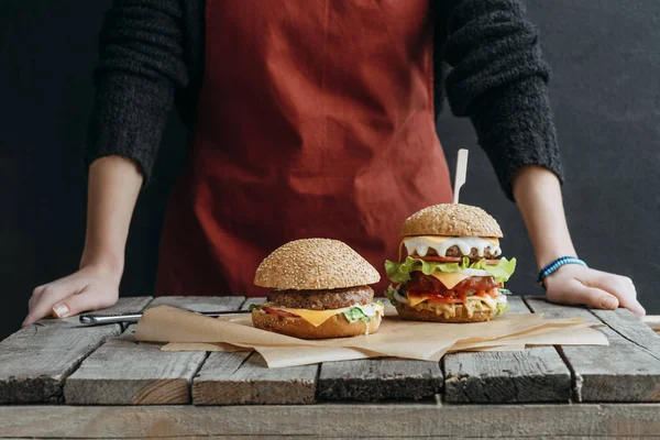 Cropped view of girl in apron standing at wooden table with tasty hamburgers on baking paper — Stock Photo