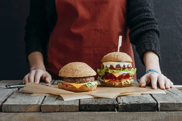 Vista recortada de la chica en delantal de pie en la mesa de madera con sabrosas hamburguesas con queso sobre papel de hornear — Stock Photo