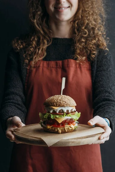 Vista recortada de chica en delantal sosteniendo tablero de madera con hamburguesa de queso casera grande - foto de stock