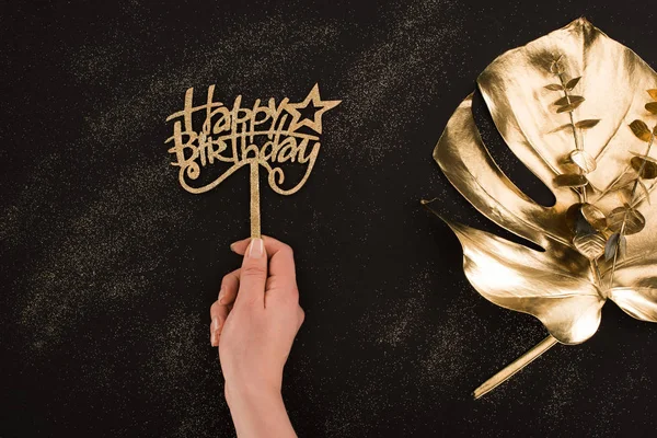 Recortado disparo de la mano femenina sosteniendo feliz cumpleaños signo con hoja de oro que se encuentra cerca de - foto de stock