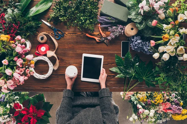 Cropped image of florist holding coffee in paper cup and tablet in hands — Stock Photo