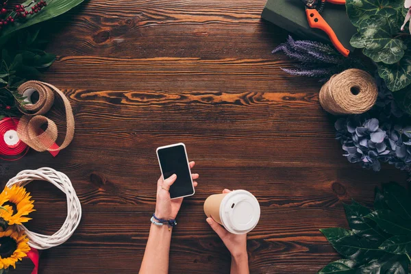Image recadrée de fleuriste tenant tasse de café jetable et smartphone dans les mains — Photo de stock