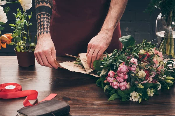 Cropped image of male florist with tattoo on hand wrapping bouquet in pack paper — Stock Photo